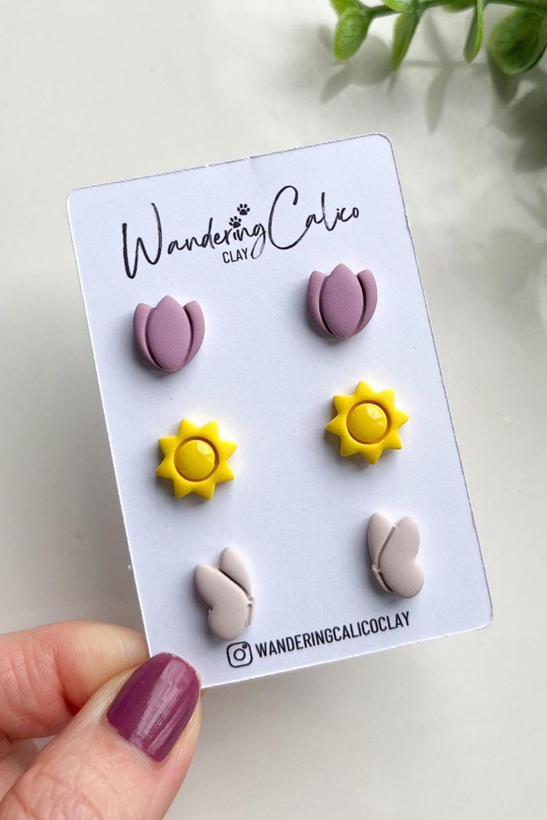 Puede incluir: Un conjunto de seis pendientes de arcilla polim&eacute;rica con tres dise&ntilde;os diferentes: una flor rosa, un sol amarillo y una mariposa blanca. Los pendientes se muestran en una tarjeta blanca con el texto "Wandering Calico Clay" y un nombre de usuario de Instagram.