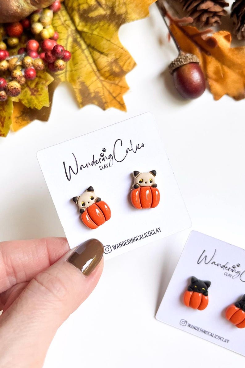 May include: A pair of cat-shaped earrings nestled in orange pumpkins, displayed on a white card. The earrings are made of clay and feature a black and white cat face. The card has the text "Wandering Calico Clay" and the Instagram handle.