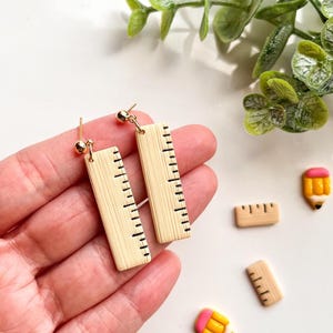 May include: A pair of wooden ruler earrings with gold hooks. The earrings are in the shape of a ruler with black markings. The earrings are being held in a hand.