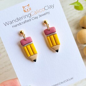 May include: A pair of handcrafted clay earrings shaped like yellow pencils with pink erasers and silver accents. The earrings have gold-colored posts and are displayed on a white card with the text "WanderingCalicoClay".
