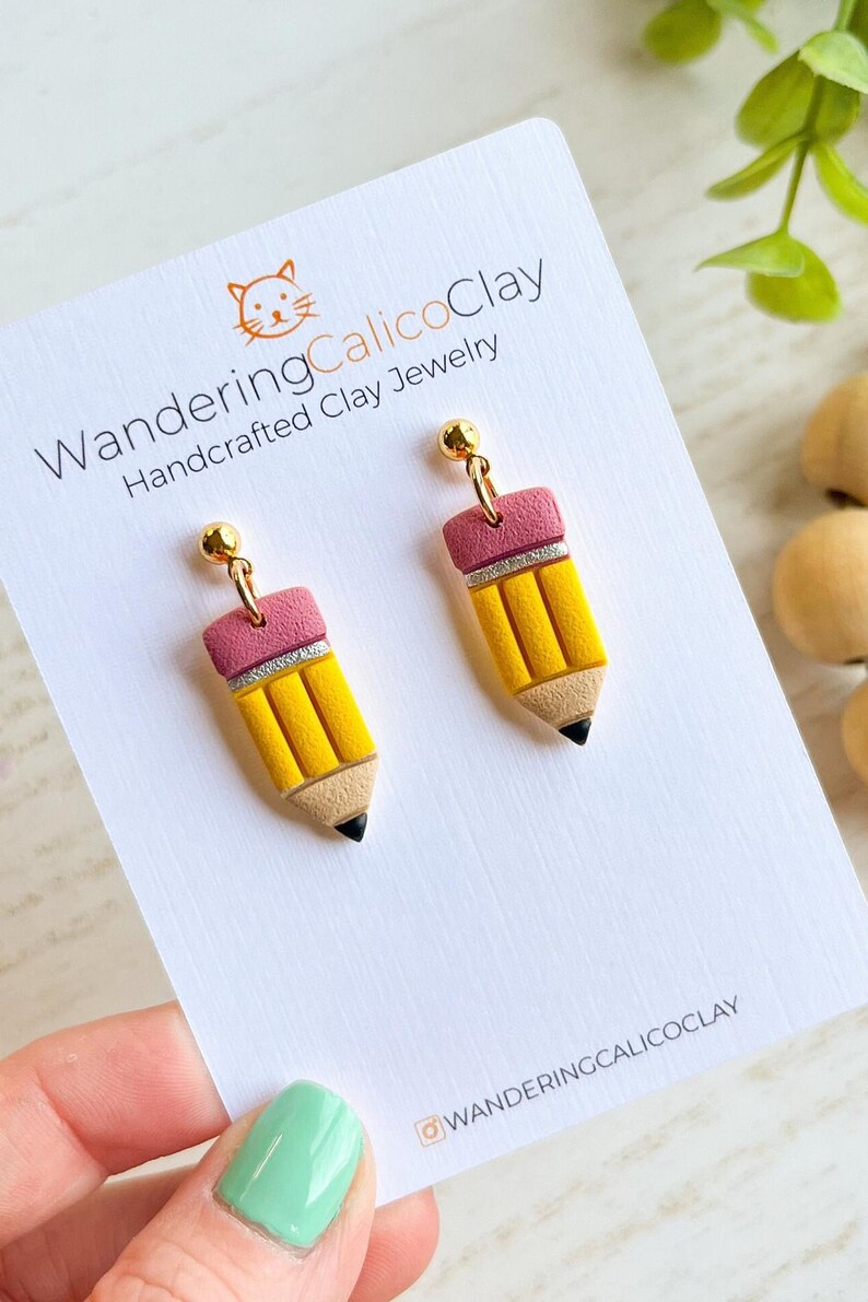 May include: A pair of handcrafted clay earrings shaped like yellow pencils with pink erasers and silver accents. The earrings have gold-colored posts and are displayed on a white card with the text "WanderingCalicoClay".