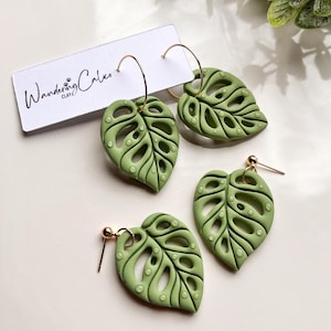 Puede incluir: Un conjunto de tres pendientes de hoja de monstera verde. Los pendientes están hechos de arcilla polimérica y tienen un acabado dorado. El pendiente más grande es un pendiente de aro, mientras que los otros dos son pendientes colgantes.