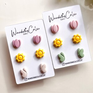 Puede incluir: Dos juegos de pendientes de arcilla polim&eacute;rica. El primer juego presenta flores rosas, soles amarillos y mariposas rosas. El segundo juego presenta flores rosas, soles amarillos y mariposas verdes. Ambos juegos tienen el texto "Wandering Calico Clay" en las tarjetas.