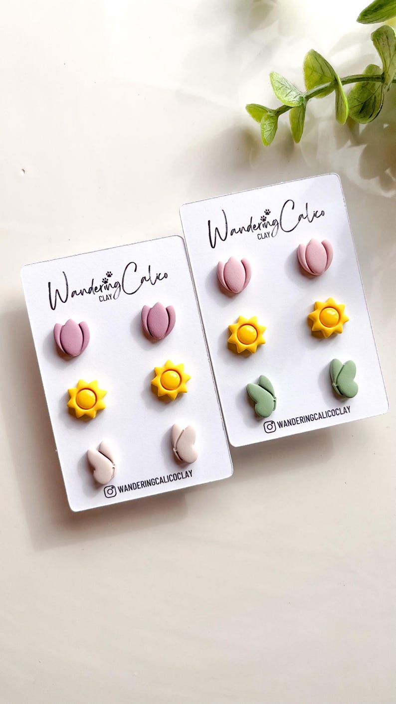 Puede incluir: Dos juegos de pendientes de arcilla polim&eacute;rica. El primer juego presenta flores rosas, soles amarillos y mariposas rosas. El segundo juego presenta flores rosas, soles amarillos y mariposas verdes. Ambos juegos tienen el texto "Wandering Calico Clay" en las tarjetas.