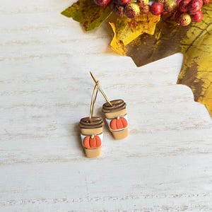 May include: A pair of gold hoop earrings featuring miniature coffee cup charms. Each charm is tan with a brown lid and a carved orange pumpkin. The earrings are set against a white wooden background with fall foliage.