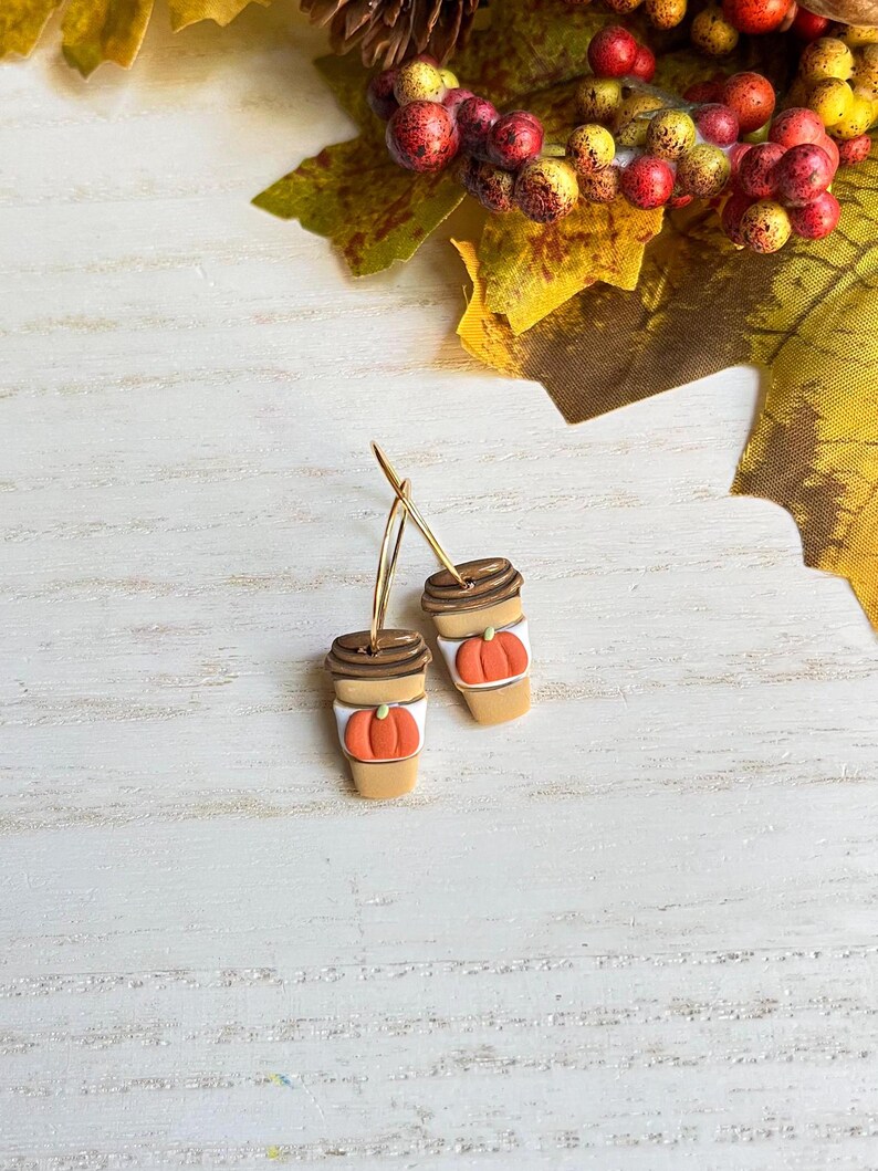 May include: A pair of gold hoop earrings featuring miniature coffee cup charms. Each charm is tan with a brown lid and a carved orange pumpkin. The earrings are set against a white wooden background with fall foliage.