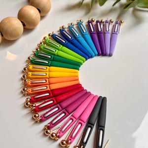 May include: A vibrant collection of colorful pen-shaped charms arranged in a semi-circle. The charms are in a rainbow of colors, including red, orange, yellow, green, blue, and purple. Each charm has a gold-colored top and a silver-colored pen tip.