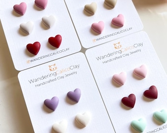 May include: Set of six pairs of handmade heart-shaped clay earrings in various colours, including pink, red, white, purple, and green. The earrings are displayed on white cards with the brand name "Wandering CalicoClay" and the social media handle "@wanderingcalicoClay".
