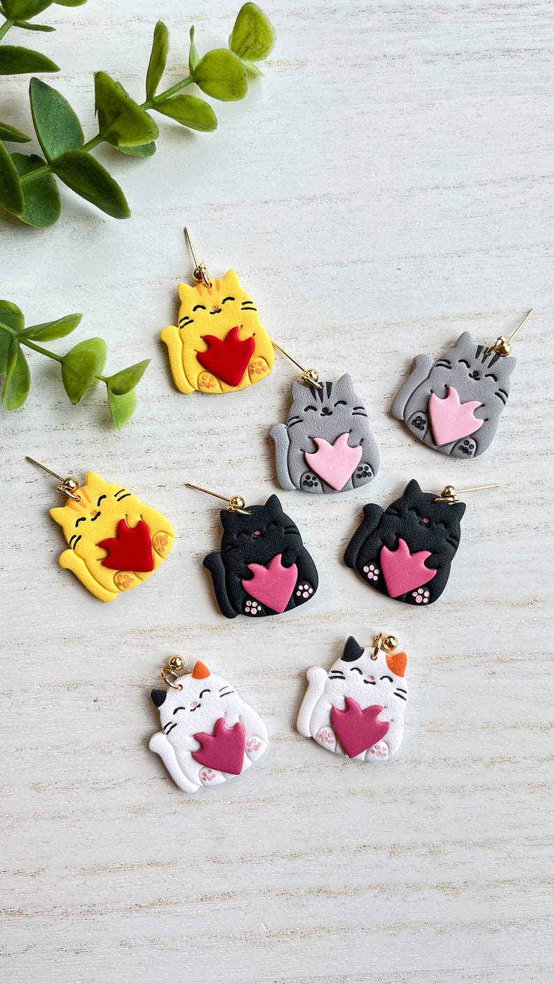 May include: Six pairs of cat-shaped earrings with a heart design. The cats are in various colors, including yellow, gray, black, and white. Each cat is holding a heart in its paws.