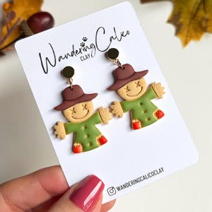 May include: A pair of dangle earrings featuring clay scarecrows with brown hats, green shirts, and orange patches. The earrings are attached to a white display card with the text Wandering Calico and WANDERINGCALICOCLAY.