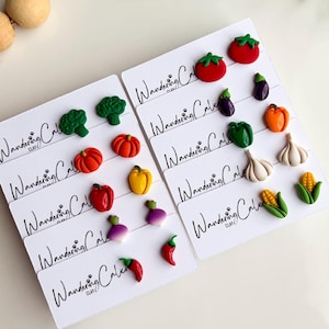 Könnte beinhalten: Eine Sammlung farbenfroher, miniaturisierter Ohrringe in Gemüseform, darunter Tomaten, Brokkoli und Paprika. Die Ohrringe werden auf weißen Karten mit dem Text "Wandering Clay" in schwarzer Schrift präsentiert.