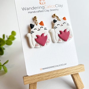 May include: A pair of white cat-shaped earrings with red hearts, holding the hearts in their paws. The earrings are handcrafted clay jewelry by WanderingCalicoClay.
