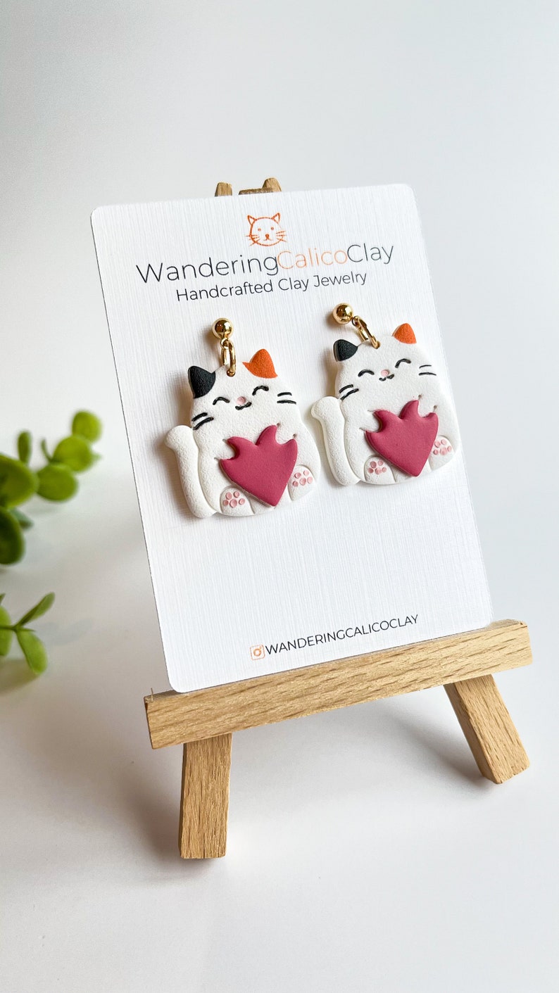 May include: A pair of white cat-shaped earrings with red hearts, holding the hearts in their paws. The earrings are handcrafted clay jewelry by WanderingCalicoClay.
