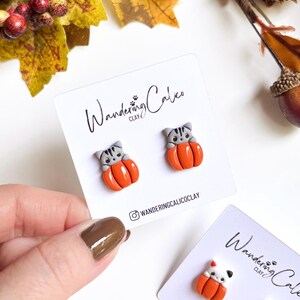 May include: A pair of cat-shaped earrings nestled in orange pumpkins, displayed on a white card. The earrings are gray and orange, with the brand name "Wandering Calico Clay" printed on the card. The background features fall foliage.