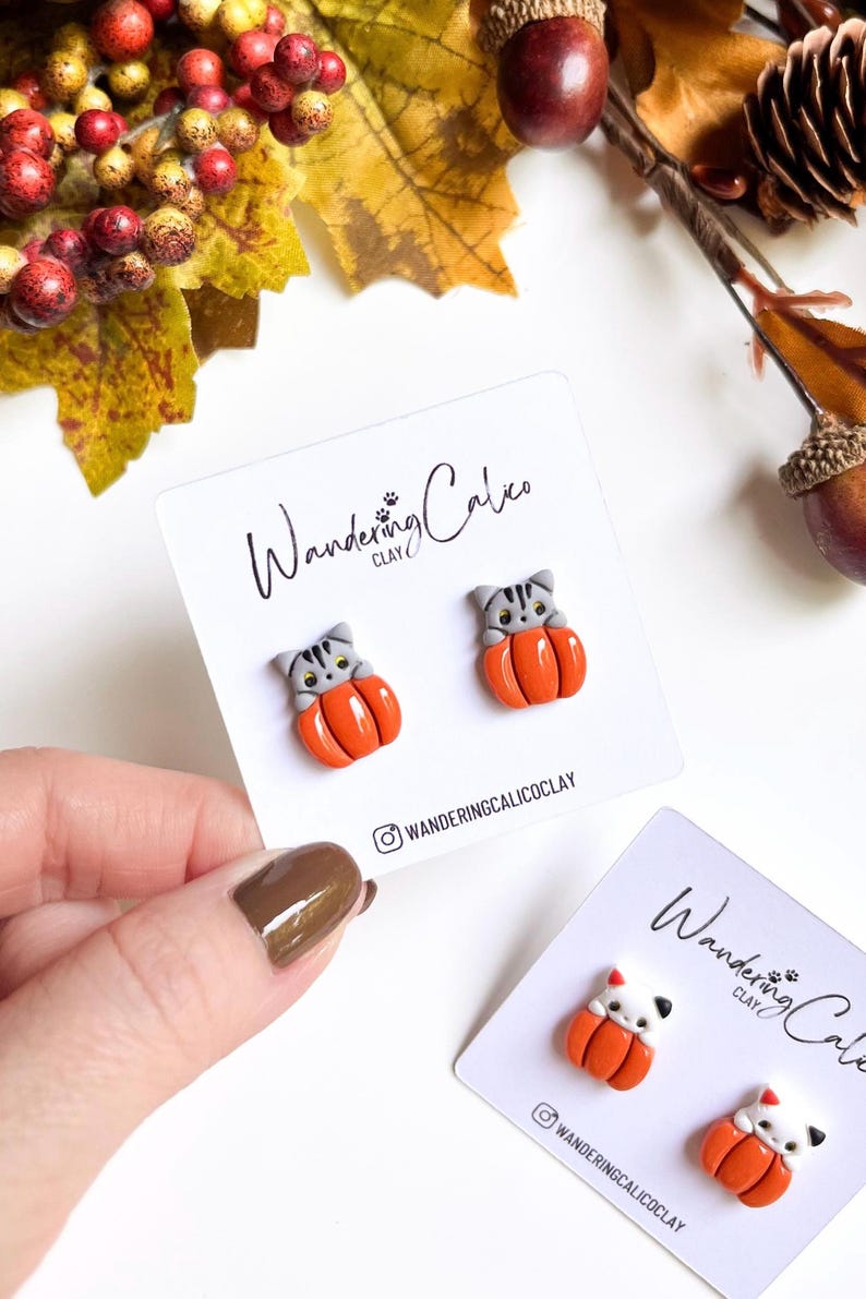 May include: A pair of cat-shaped earrings nestled in orange pumpkins, displayed on a white card. The earrings are gray and orange, with the brand name "Wandering Calico Clay" printed on the card. The background features fall foliage.