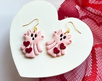 May include: Pink ghost-shaped earrings with red hearts. The earrings are hanging from a white heart-shaped surface.