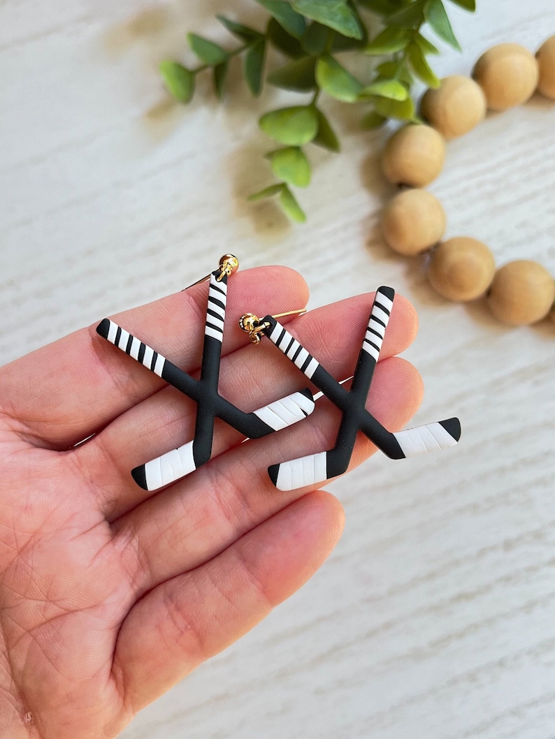 K&ouml;nnte beinhalten: Ein Paar schwarze und wei&szlig;e Hockey-Schl&auml;ger-Ohrringe mit goldenen Akzenten. Die Ohrringe sind aus Polymer Clay gefertigt und sollen wie Hockeyschl&auml;ger aussehen.