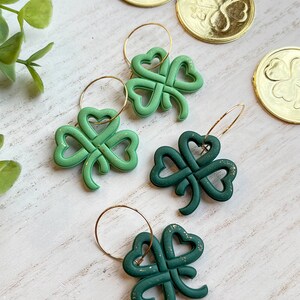 May include: Two pairs of green shamrock earrings with gold glitter accents. The earrings are on a white wooden surface with gold coins featuring shamrock designs.