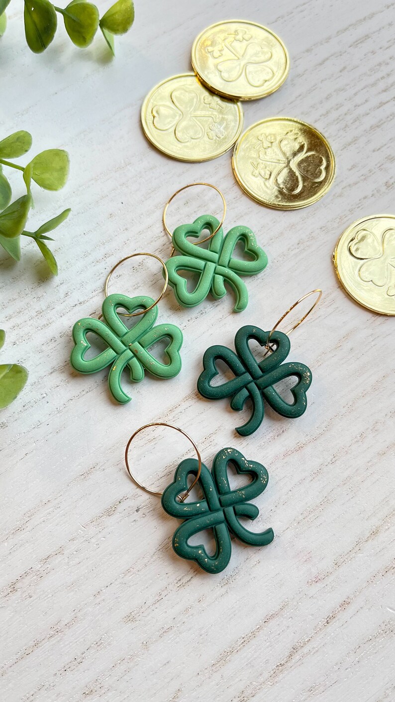 May include: Two pairs of green shamrock earrings with gold glitter accents. The earrings are on a white wooden surface with gold coins featuring shamrock designs.