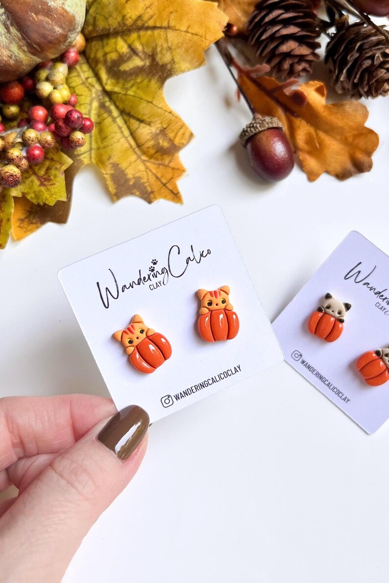 May include: Orange and tan cat-shaped earrings on white cards. The earrings are shaped like pumpkins with cat faces. The cards have the text "Wandering Calico Clay" and an Instagram handle. Autumn leaves and acorns are in the background.