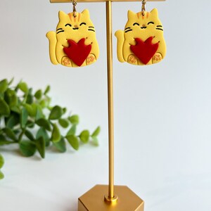 May include: A pair of yellow cat earrings with red hearts. The earrings are on a gold stand.