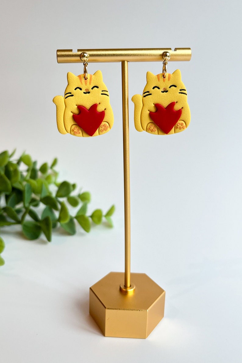 May include: A pair of yellow cat earrings with red hearts. The earrings are on a gold stand.