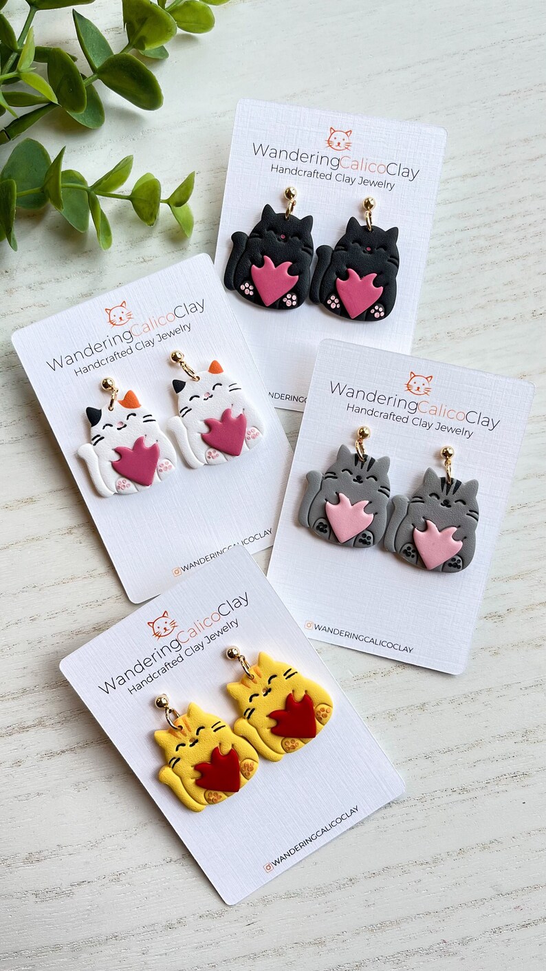 May include: Three pairs of handmade polymer clay cat earrings. Each pair features a cat holding a heart in its paws. The cats are in black, white, and yellow.