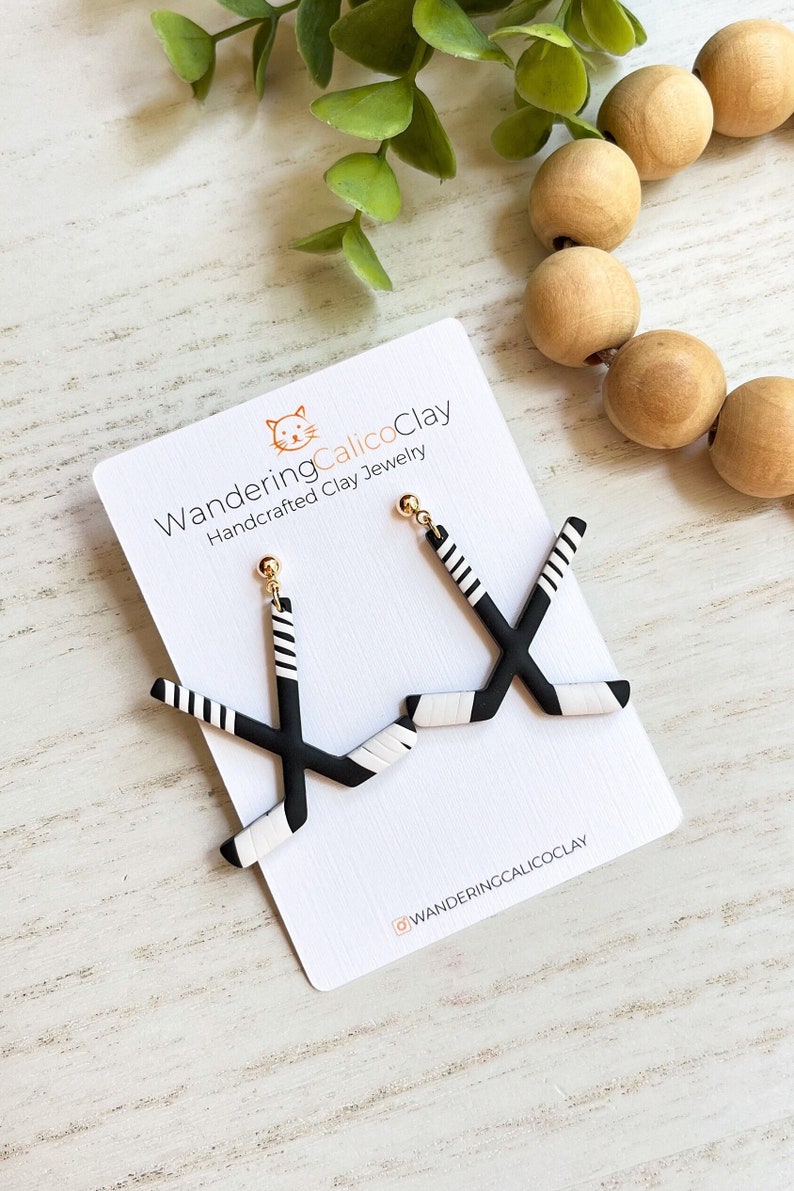 K&ouml;nnte beinhalten: Schwarze und wei&szlig;e Hockey-Schl&auml;ger-Ohrringe mit goldenen Haken. Die Ohrringe befinden sich auf einer wei&szlig;en Karte mit dem Text "WanderingCalicoClay Handcrafted Clay Jewelry" und dem Instagram-Handle "@wanderingcalicoclay".