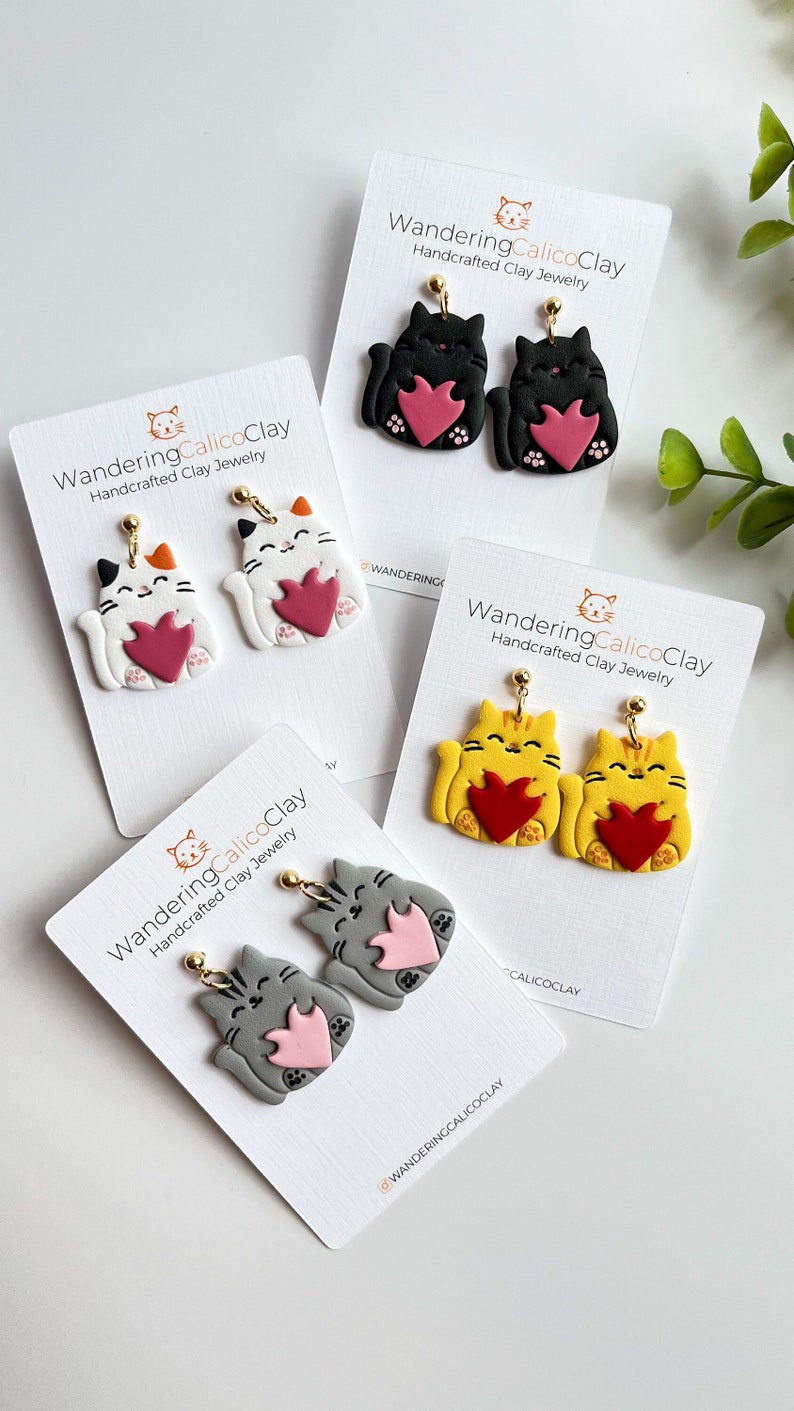 May include: Four pairs of cat-shaped polymer clay earrings. Each pair features a cat holding a heart in its paws. The cats are in different colors: black, white, yellow, and gray. The earrings are handcrafted and have gold-colored hooks.