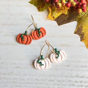 May include: A pair of orange pumpkin earrings with green stems and gold hoops, and a pair of white pumpkin earrings with green stems and gold hoops. The earrings are set against a white wooden background with fall foliage.