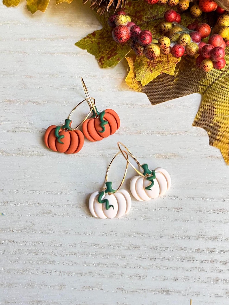 May include: A pair of orange pumpkin earrings with green stems and gold hoops, and a pair of white pumpkin earrings with green stems and gold hoops. The earrings are set against a white wooden background with fall foliage.