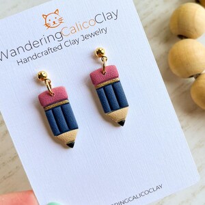 May include: A pair of handcrafted clay earrings shaped like pencils. The earrings feature a blue pencil body, a pink eraser, and a gold band. The earrings are attached to gold-colored ball studs. The earrings are displayed on a white card with the text "WanderingCalicoClay".
