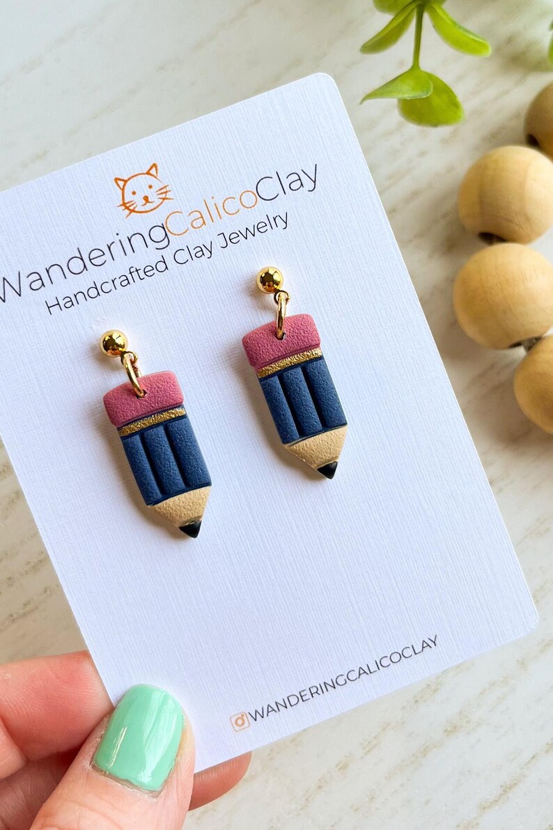 May include: A pair of handcrafted clay earrings shaped like pencils. The earrings feature a blue pencil body, a pink eraser, and a gold band. The earrings are attached to gold-colored ball studs. The earrings are displayed on a white card with the text "WanderingCalicoClay".