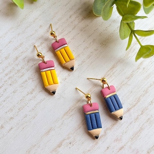 May include: Earrings shaped like pencils, featuring yellow, pink, and blue designs. The pencil earrings have gold-colored ear wires and black tips. The earrings are a fun accessory for anyone who loves school supplies or unique jewelry.