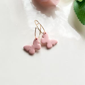 May include: A pair of pink butterfly earrings with gold hoops. The butterflies have a textured surface and are attached to the hoops with a small loop.