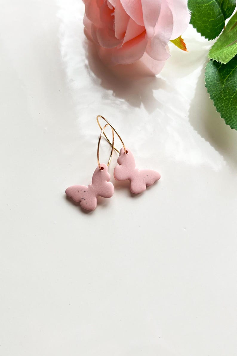 May include: A pair of pink butterfly earrings with gold hoops. The butterflies have a textured surface and are attached to the hoops with a small loop.