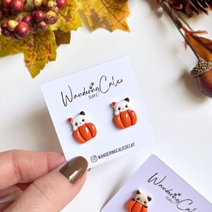 May include: A pair of handmade cat-shaped earrings, each perched atop an orange pumpkin. The earrings are displayed on a white card with the text "Wandering Calico Clay" and the Instagram handle. Autumn-themed jewelry.