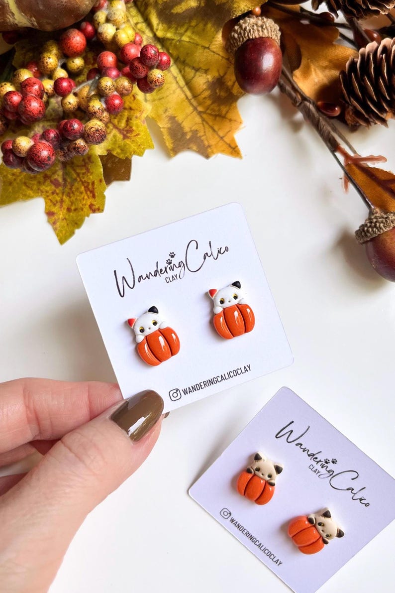May include: A pair of handmade cat-shaped earrings, each perched atop an orange pumpkin. The earrings are displayed on a white card with the text "Wandering Calico Clay" and the Instagram handle. Autumn-themed jewelry.