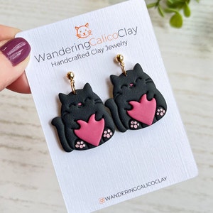 May include: A pair of black cat earrings with pink hearts. The earrings are handmade and feature gold hooks. The earrings are on a white card with the text "WanderingCalicoClay Handcrafted Clay Jewelry" and "@WanderingCalicoClay".