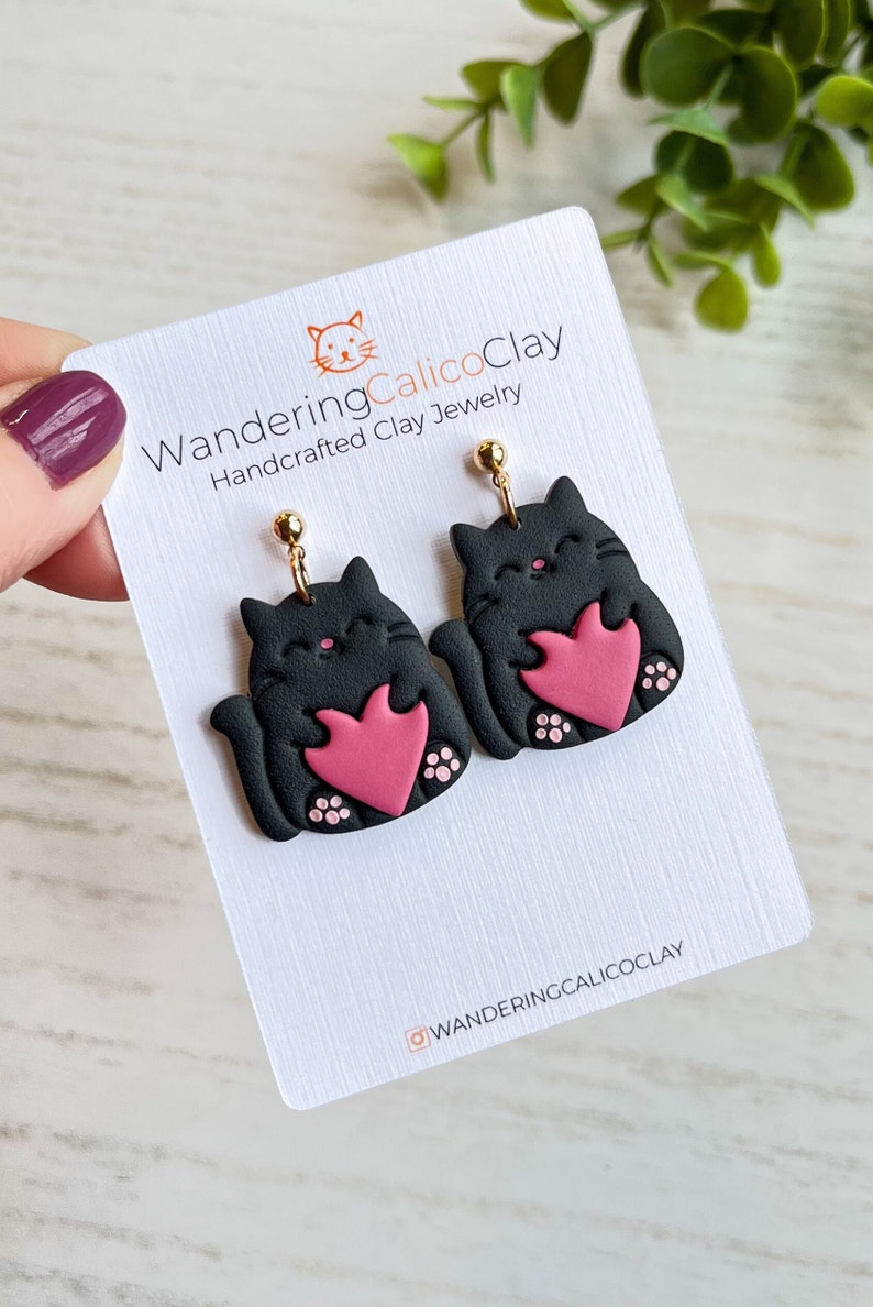 May include: A pair of black cat earrings with pink hearts. The earrings are handmade and feature gold hooks. The earrings are on a white card with the text "WanderingCalicoClay Handcrafted Clay Jewelry" and "@WanderingCalicoClay".