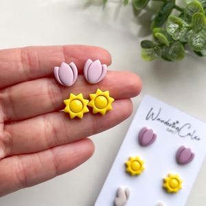 Puede incluir: Un conjunto de tres pares de pendientes de arcilla polim&eacute;rica. Los pendientes tienen forma de tulipanes y soles. Los tulipanes son de color rosa claro y los soles son amarillos. Los pendientes se muestran en una tarjeta blanca con el texto "Wandering Clay" impreso en ella.