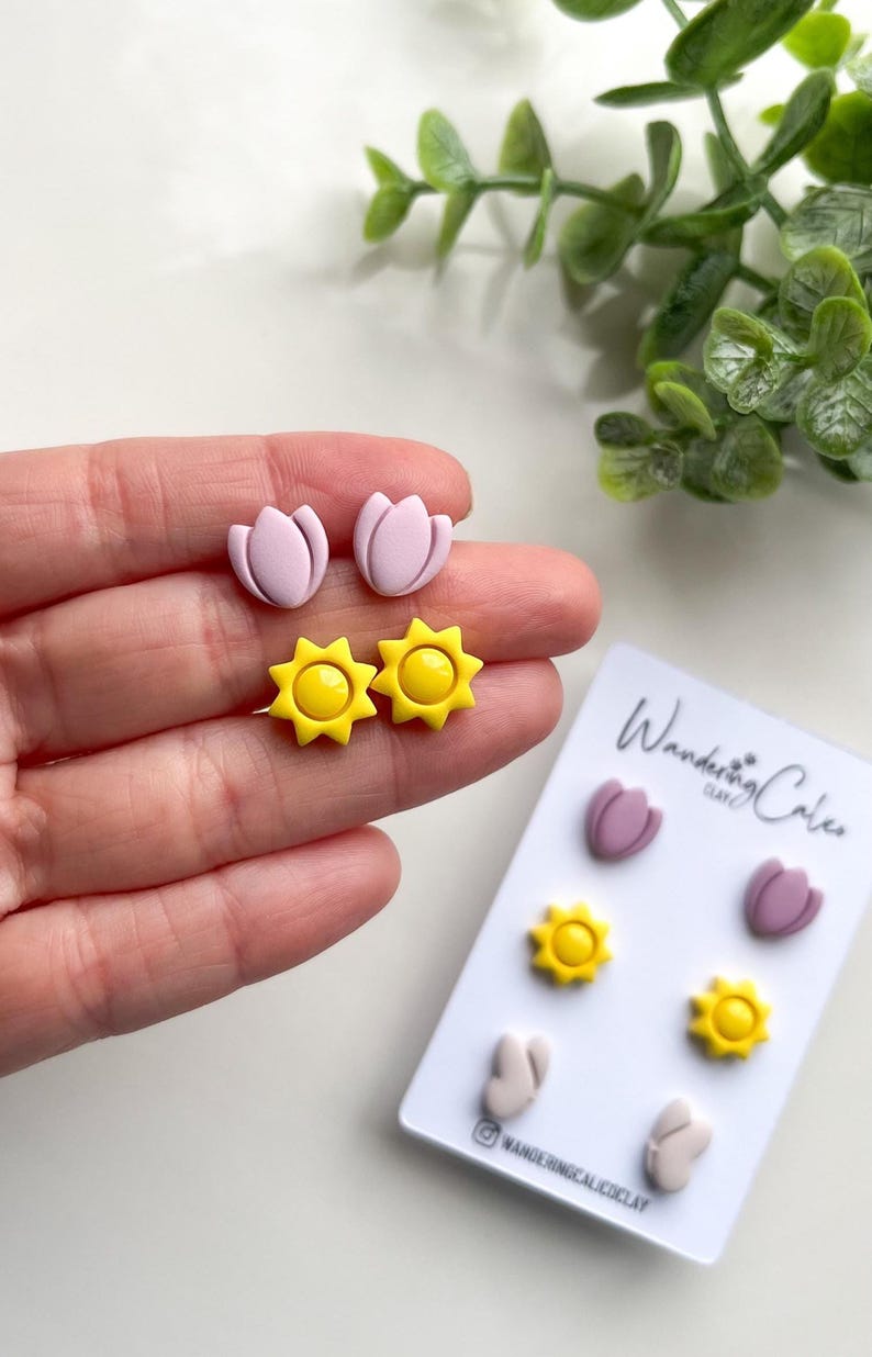 Puede incluir: Un conjunto de tres pares de pendientes de arcilla polim&eacute;rica. Los pendientes tienen forma de tulipanes y soles. Los tulipanes son de color rosa claro y los soles son amarillos. Los pendientes se muestran en una tarjeta blanca con el texto "Wandering Clay" impreso en ella.