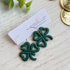 May include: A pair of green four-leaf clover earrings with gold glitter accents. The earrings are made of polymer clay and are hanging from gold hoops. The earrings are on a white wooden surface with gold coins in the background. The text "WanderingCalicoClay handcrafted clay jewelry @WANDERINGCALICOCLAY" is on a white card behind the earrings.