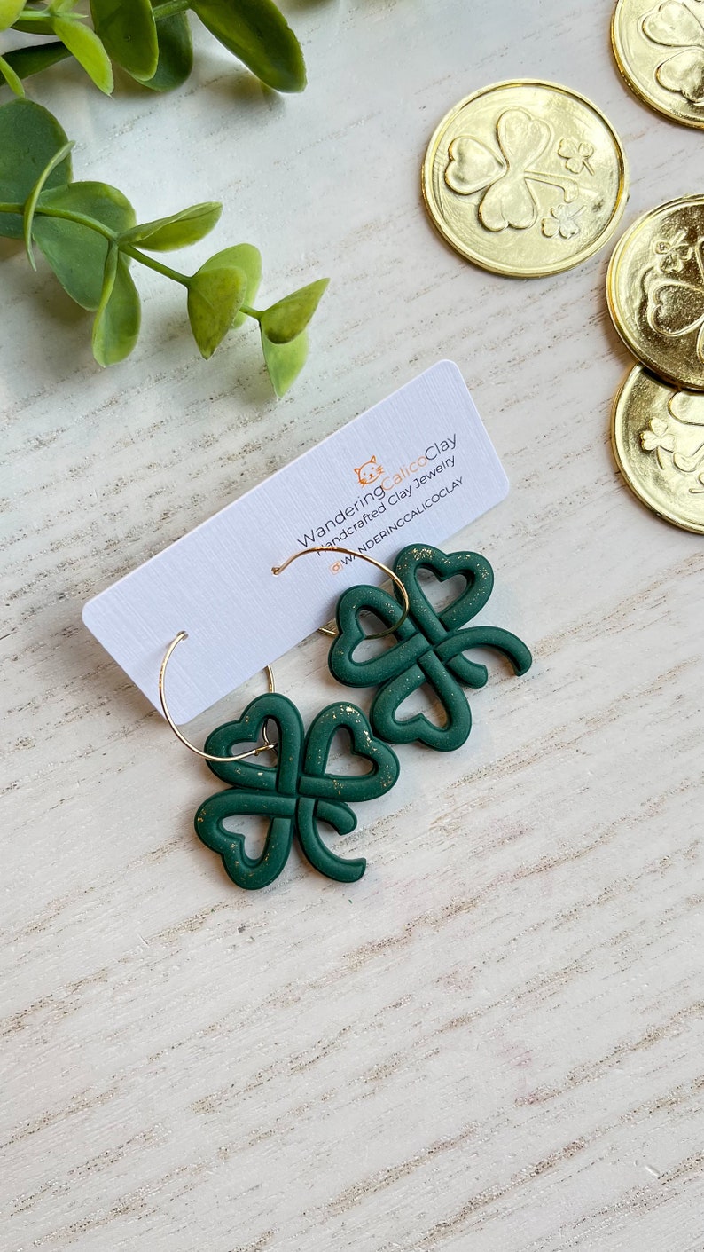 May include: A pair of green four-leaf clover earrings with gold glitter accents. The earrings are made of polymer clay and are hanging from gold hoops. The earrings are on a white wooden surface with gold coins in the background. The text "WanderingCalicoClay handcrafted clay jewelry @WANDERINGCALICOCLAY" is on a white card behind the earrings.