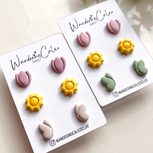 Puede incluir: Dos juegos de pendientes de arcilla polim&eacute;rica en tarjetas blancas. El primer juego presenta tulipanes rosas, soles amarillos y mariposas beige. El segundo juego presenta tulipanes rosas, soles amarillos y mariposas verdes. Las tarjetas tienen el texto "Wandering Calico Clay" impreso en ellas.