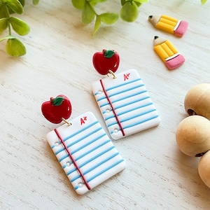 May include: A pair of earrings featuring a red apple and a white sheet of paper with blue lines and a red "A+" grade. The paper has a red border and a row of white dots along the edge.