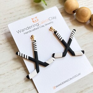 K&ouml;nnte beinhalten: Ein Paar schwarze und wei&szlig;e Hockey-Schl&auml;ger-Ohrringe mit goldenen Haken. Die Ohrringe werden auf einer wei&szlig;en Karte mit dem Text "WanderingCalicoClay Handcrafted Clay Jewelry" und dem Instagram-Handle "@wanderingcalicoclay" pr&auml;sentiert.