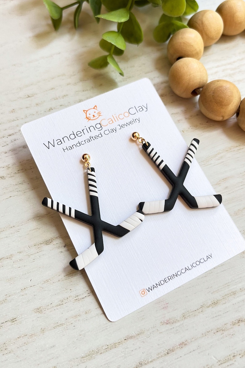 K&ouml;nnte beinhalten: Ein Paar schwarze und wei&szlig;e Hockey-Schl&auml;ger-Ohrringe mit goldenen Haken. Die Ohrringe werden auf einer wei&szlig;en Karte mit dem Text "WanderingCalicoClay Handcrafted Clay Jewelry" und dem Instagram-Handle "@wanderingcalicoclay" pr&auml;sentiert.