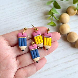 May include: Four pencil-shaped earrings in blue and yellow with pink erasers and gold-colored studs. The earrings are held in a hand against a white background with green foliage and wooden beads.