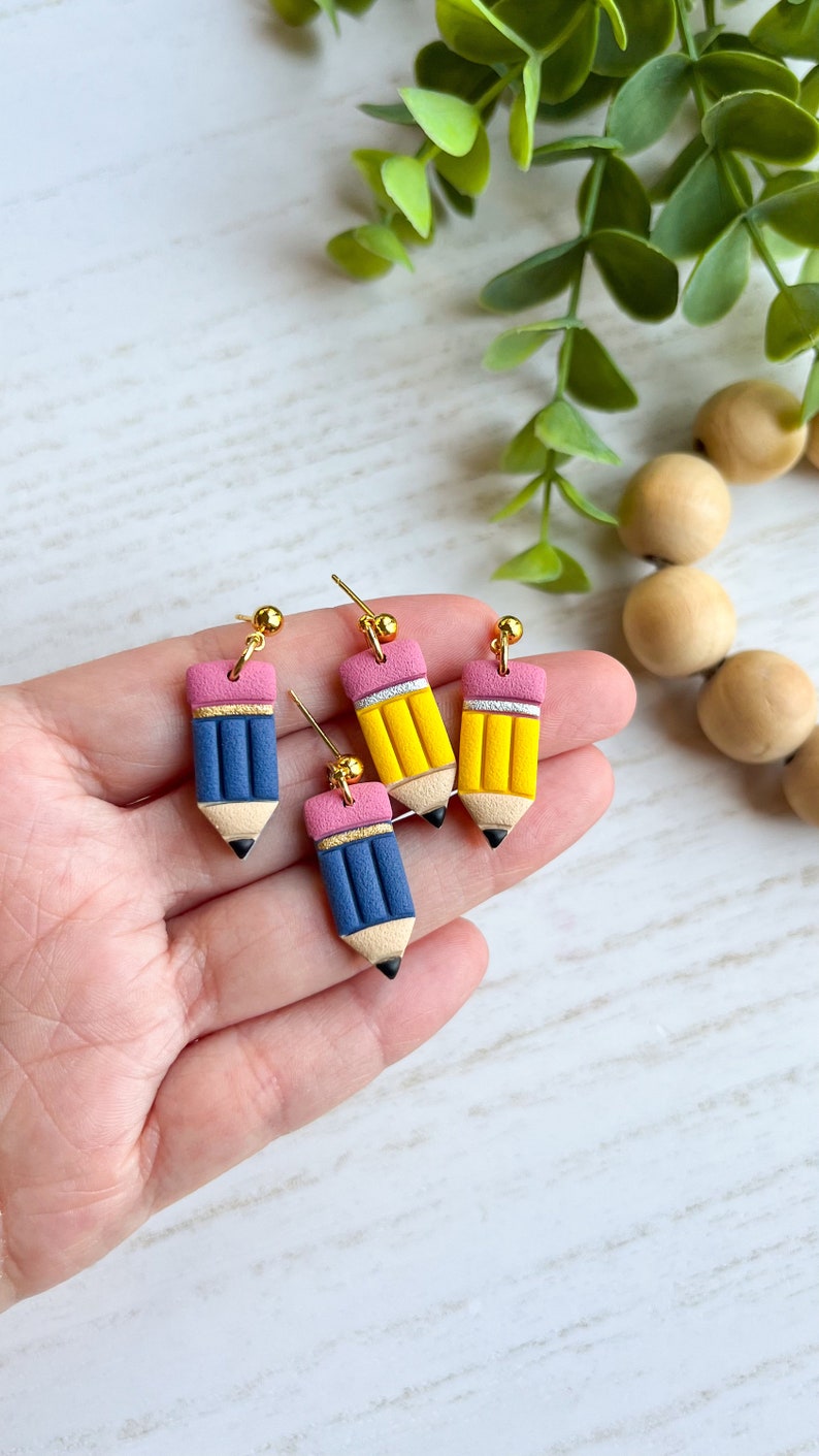 May include: Four pencil-shaped earrings in blue and yellow with pink erasers and gold-colored studs. The earrings are held in a hand against a white background with green foliage and wooden beads.